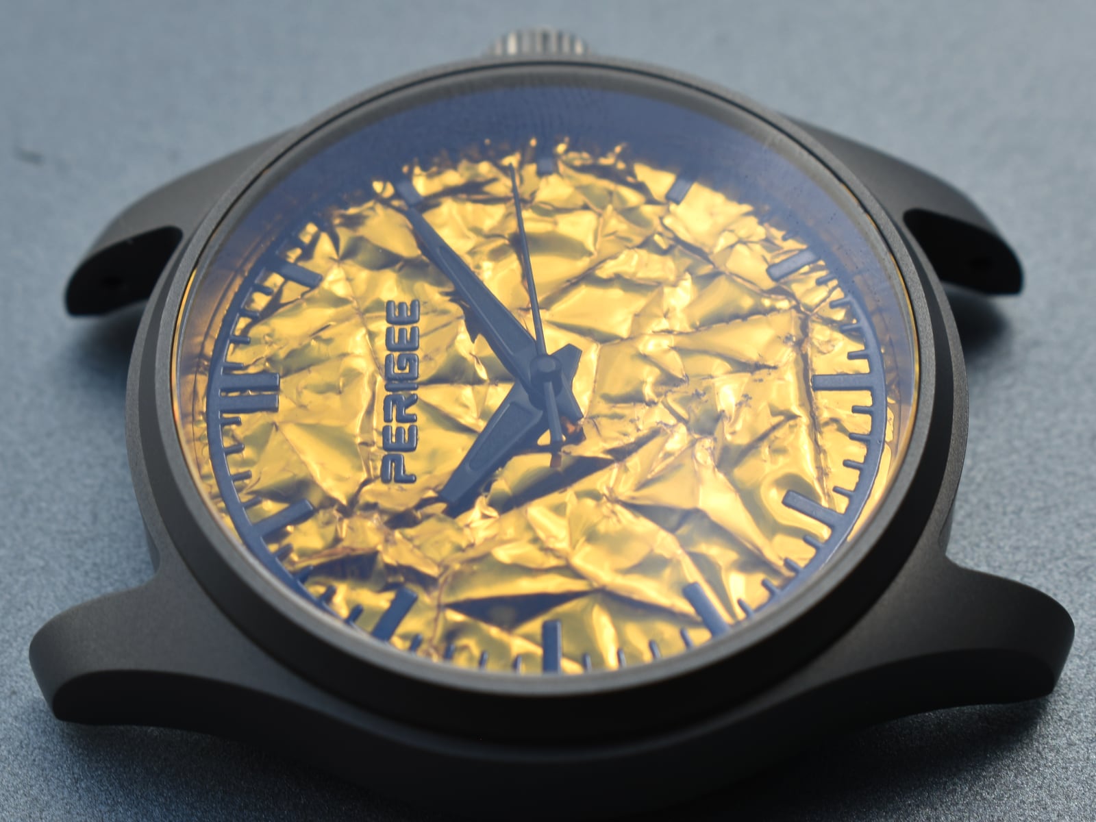 The watch at a low angle on a grey ground, showing the blasted titanium case side, the crown and the lugs.