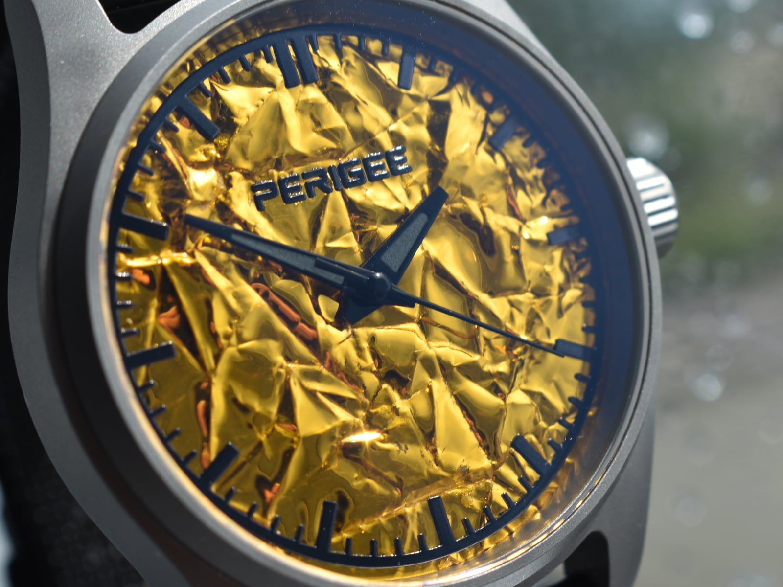The dial close in: folded aluminized foil under the hands, the PERIGEE wordmark across the upper third, every ridge catching a different amount of light.